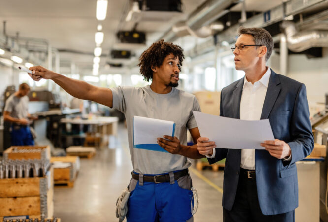 Company manager holding project plans while talking to African American manual worker who is pointing at something in the factory.