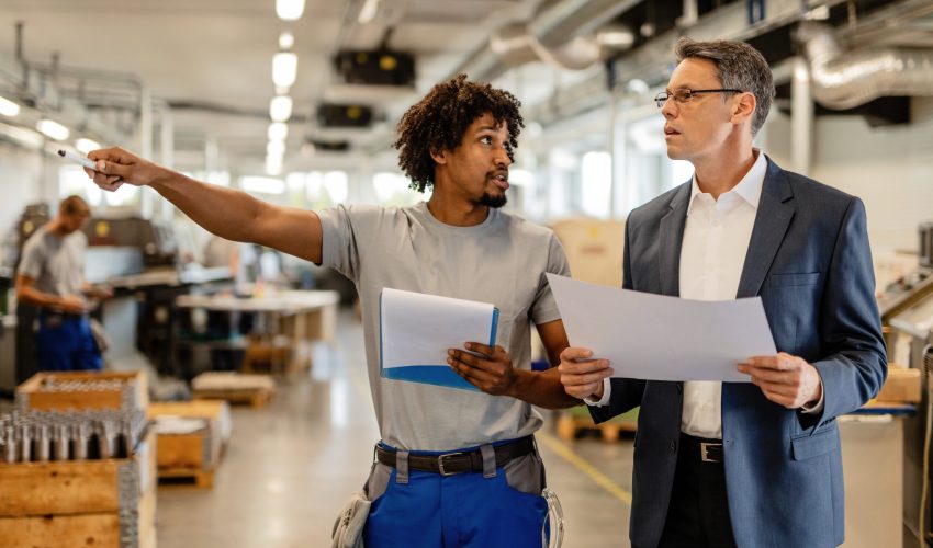 Company manager holding project plans while talking to African American manual worker who is pointing at something in the factory.