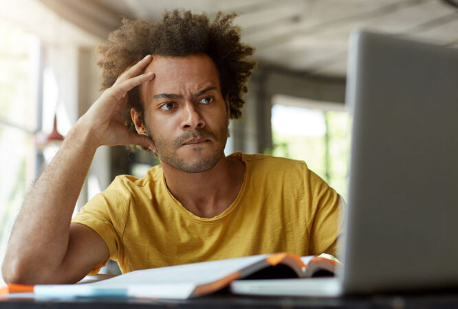 Attentive black young male sitting indoors in front of opened laptop being very serious while reading scientific article online trying to find main point of it and to write review on this topic