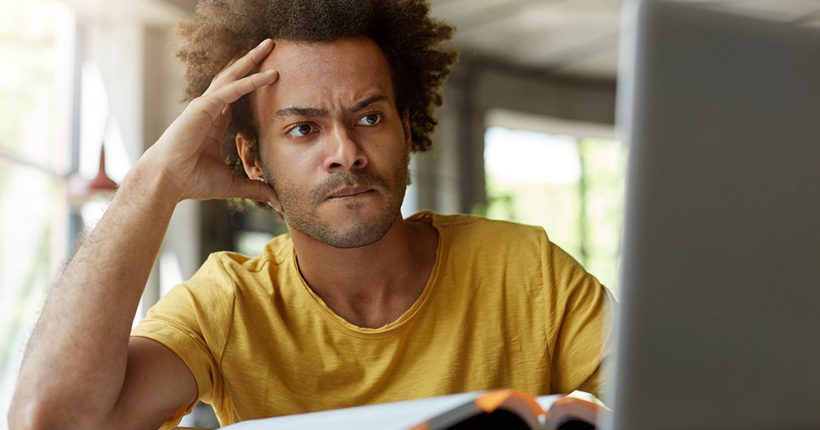 Attentive black young male sitting indoors in front of opened laptop being very serious while reading scientific article online trying to find main point of it and to write review on this topic