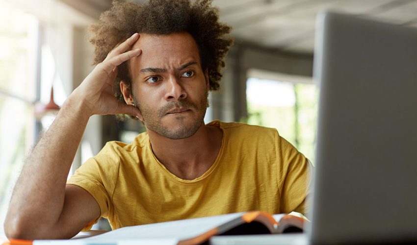 Attentive black young male sitting indoors in front of opened laptop being very serious while reading scientific article online trying to find main point of it and to write review on this topic