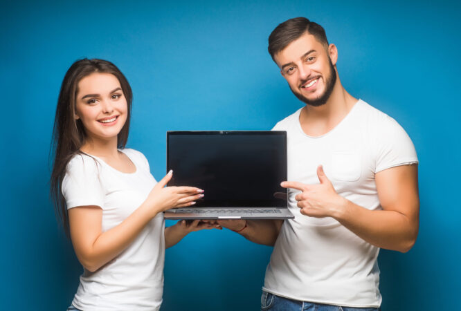 Portrait of ecstatic man and woman, happy people while holding black laptop isolated over blue background.