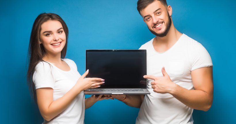 Portrait of ecstatic man and woman, happy people while holding black laptop isolated over blue background.