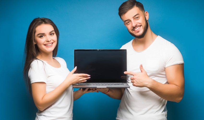 Portrait of ecstatic man and woman, happy people while holding black laptop isolated over blue background.