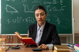 impressed young female teacher sits at table with school tools holding book and looking at calculator in her hand in classroom