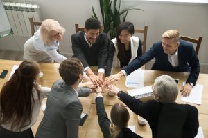 Young and old multiracial business people join pile of stacked hands together at group meeting conference table promising help support, concept of successful teamwork, team building unity, top view