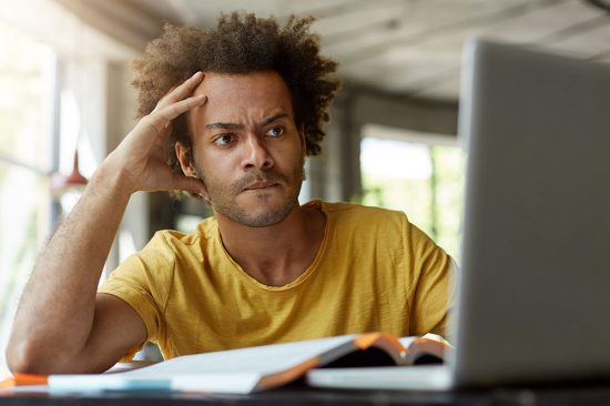 Attentive black young male sitting indoors in front of opened laptop being very serious while reading scientific article online trying to find main point of it and to write review on this topic
