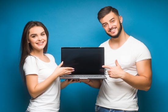 Portrait of ecstatic man and woman, happy people while holding black laptop isolated over blue background.
